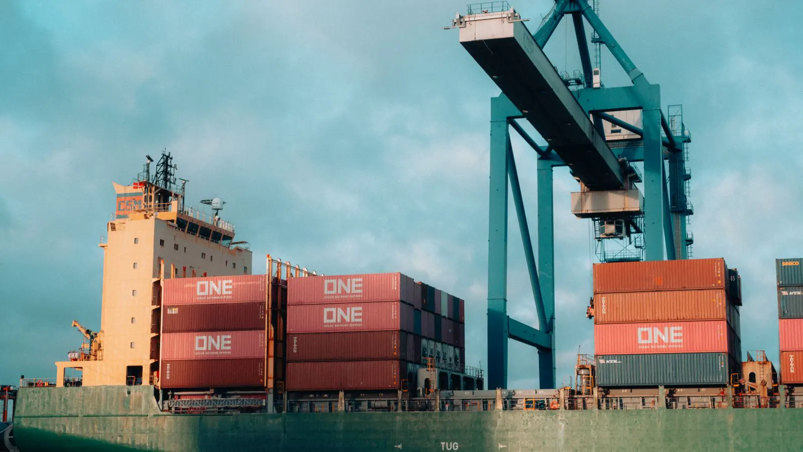 A cargo ship loading metal chemical drums on a dock.